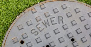 A round metal sewer cover with a textured square pattern, labeled "SEWER," is set next to green grass.