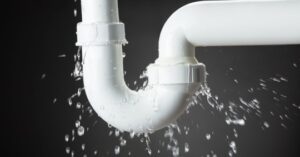 %Title% | 24/7 Rooter &Amp; Plumbing Water leaking from a white plastic pipe joint, with droplets spraying out and collecting below, against a dark background.