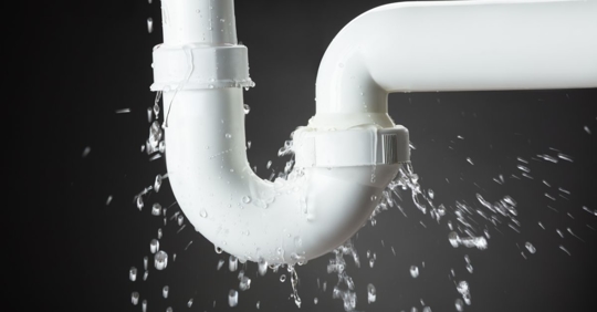 Water leaking from a white plastic pipe joint, with droplets spraying out and collecting below, against a dark background.