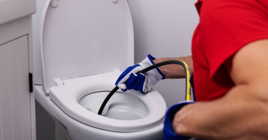 A person wearing gloves and a red shirt is using a plumbing snake to unclog a toilet.