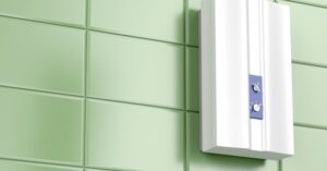 A white rectangular wall-mounted device with a blue control panel is attached to a green tiled wall.