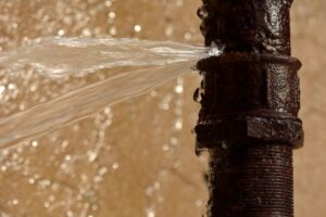 %Title% | 24/7 Rooter &Amp; Plumbing Water spraying out of a rusty, leaking pipe against a blurred background.