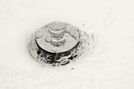 Water swirling around a metallic sink drain stopper, with bubbles and ripples visible on the surface.