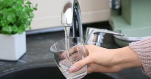 %Title% | 24/7 Rooter &Amp; Plumbing A hand holds a clear glass under a running kitchen faucet, filling it with water. A potted plant and kitchen appliances are visible in the background.