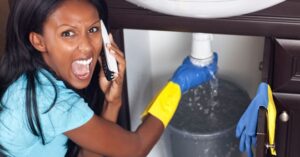 %Title% | 24/7 Rooter &Amp; Plumbing A woman on the phone appears stressed while catching water leaking from a pipe under a sink into a bucket, wearing rubber gloves.