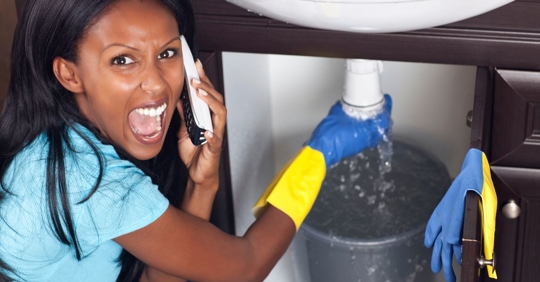 A woman on the phone appears stressed while catching water leaking from a pipe under a sink into a bucket, wearing rubber gloves.
